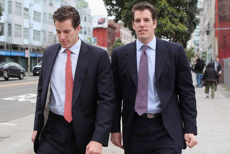 Tyler and Cameron Winklevoss are shown wearing suits, walking on a city sidewalk.